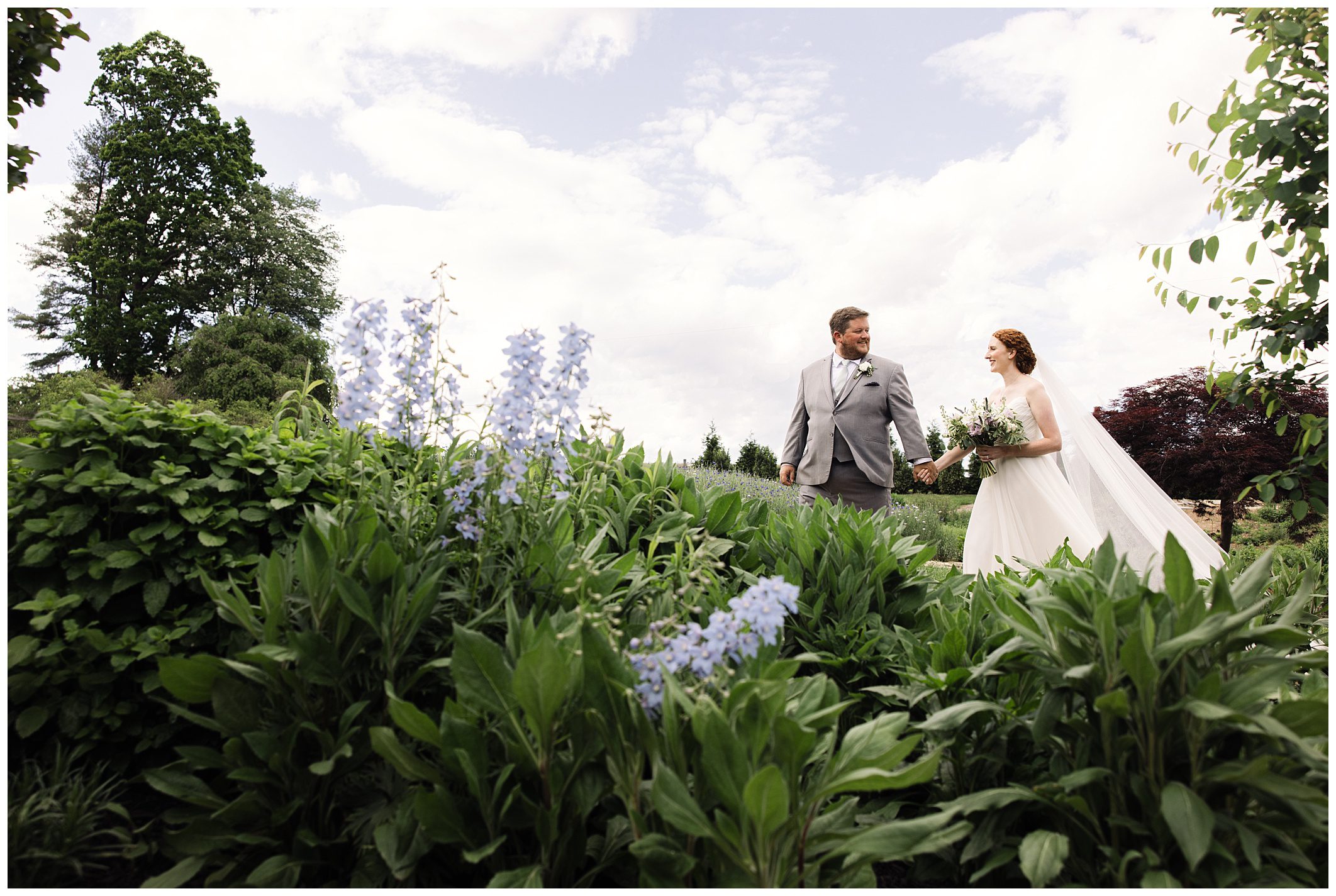 A bride and groom walk hand-in-hand through a lush garden with tall green plants and blue flowers under a partly cloudy sky.